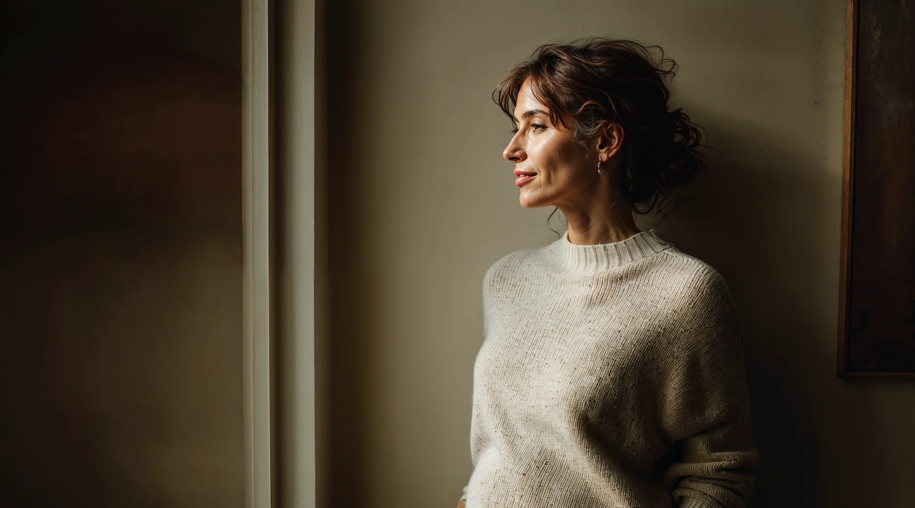 Pencere ışığında ayakta duran 46 yaş kadın, eli karın-alt bölgesinde nazik farkındalık jesti; pelvik taban farkındalığı teması