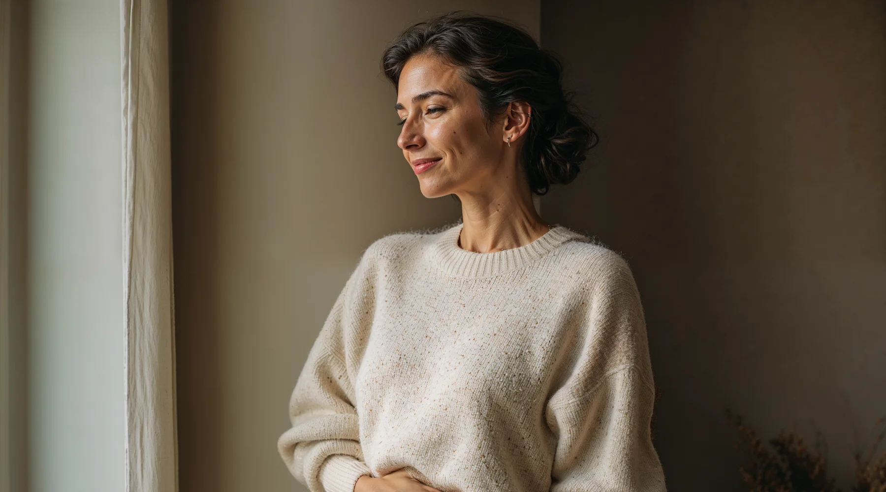 Doğal pencere ışığında kendi yanağına nazikçe dokunan 50 yaş kadın profil portresi; cilt bakımı ve öz-şefkat teması