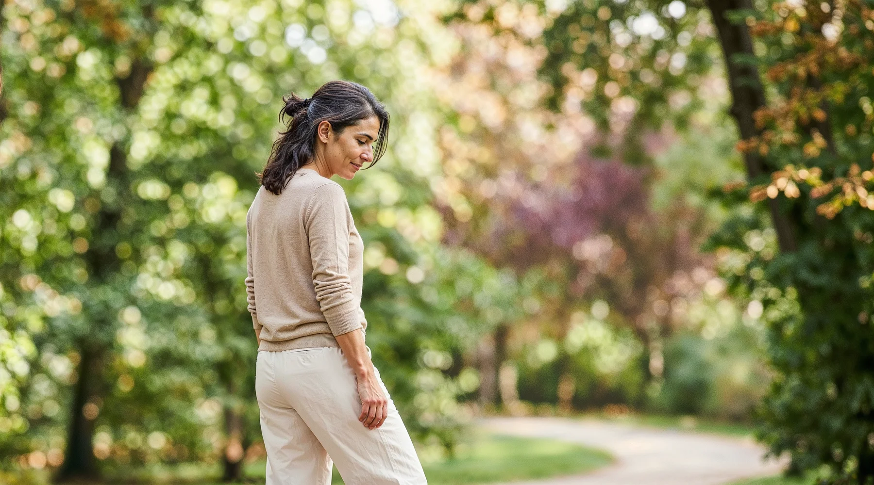 Sabah park yolunda yürüyüş molasında kadın; arkadan editöryal kadraj, krem keten pantolon, sonbahar bordo yaprak ipucu; günlük hareket ritmi teması