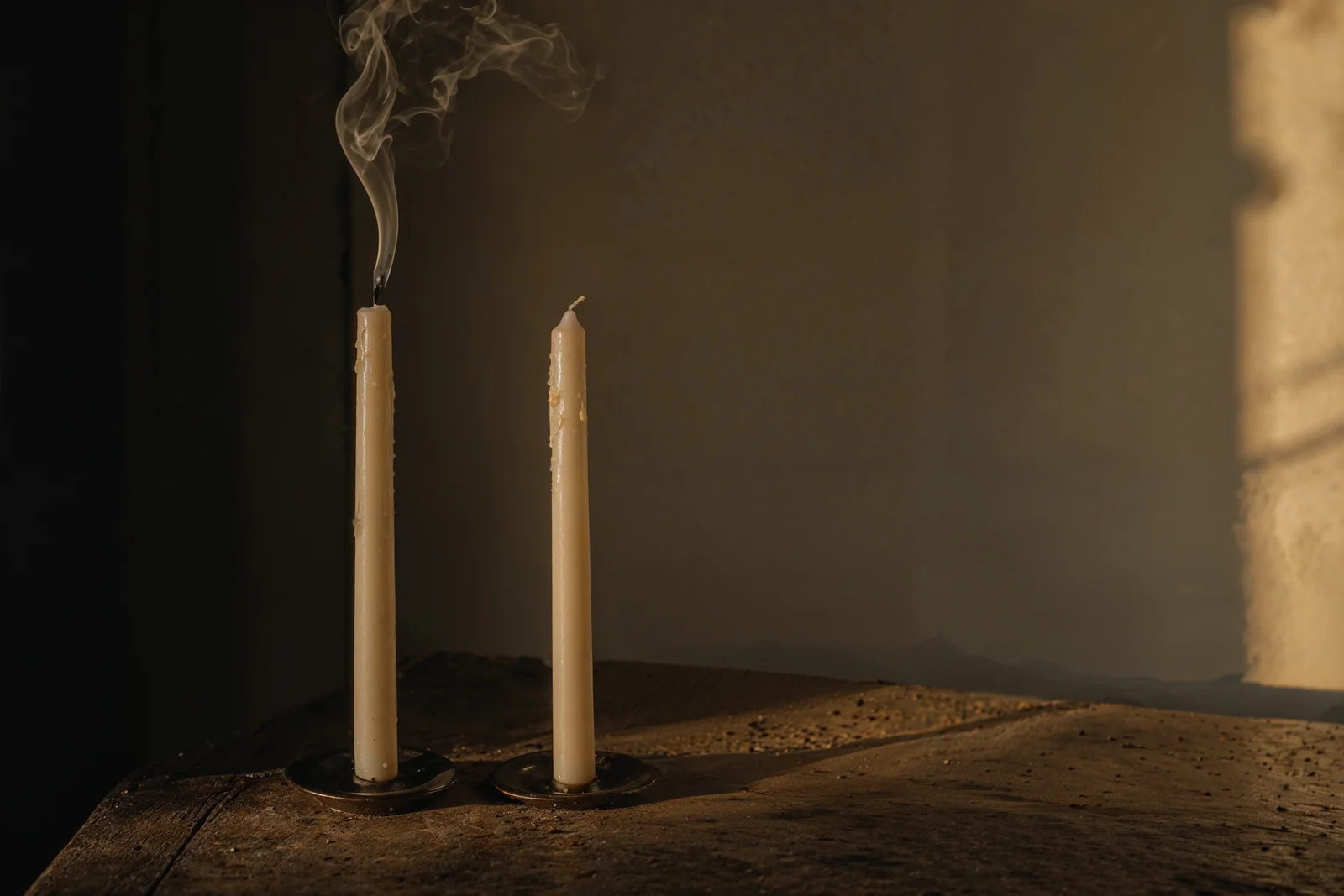 Eski ahşap masada iki uzun krem mum: biri yanmış ve tütüyor, diğeri yanmamış.