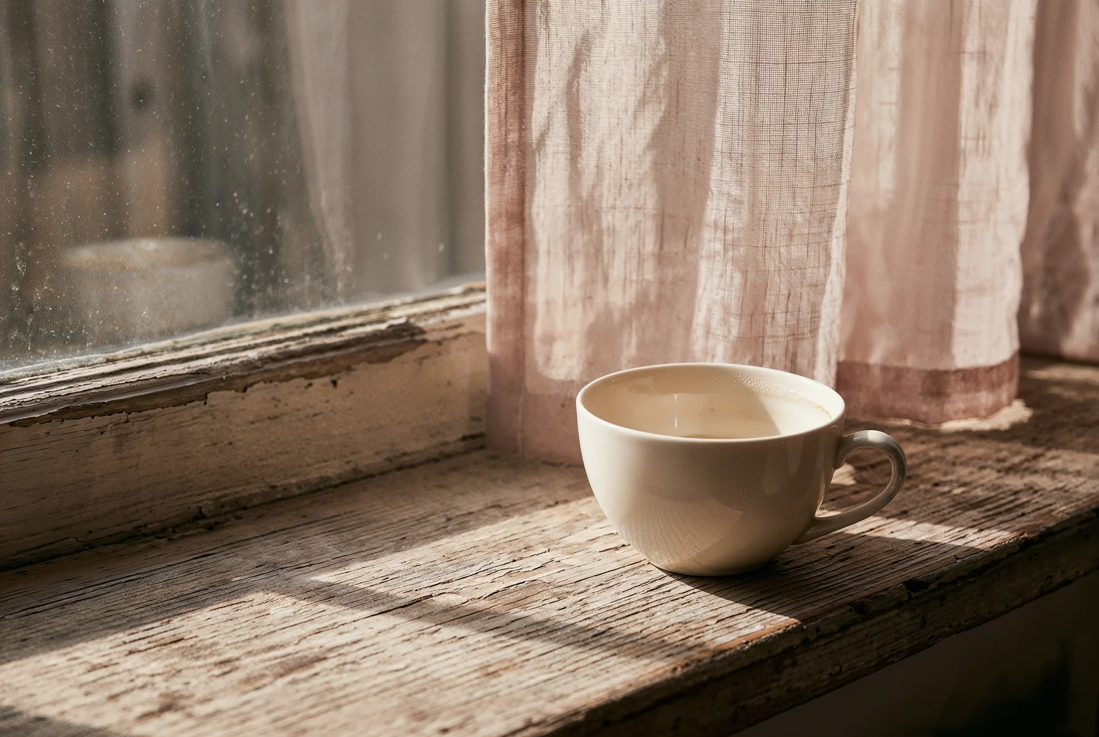 Eski pencere kenarında yarısı içilmiş porselen çay fincanı, perde gölgesi.
